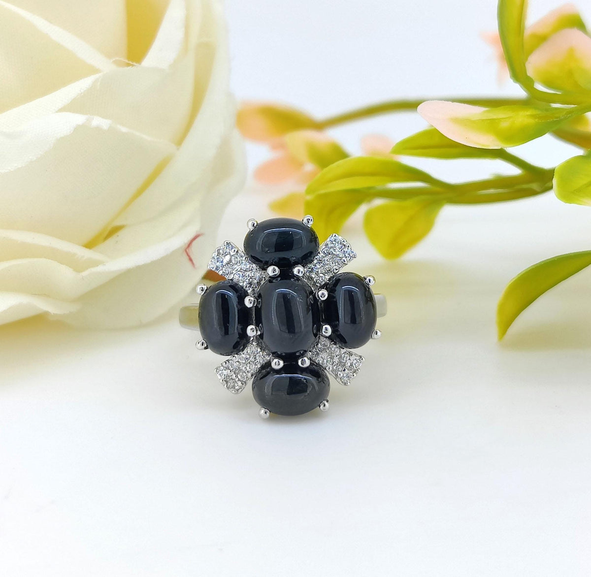 A silver ring featuring five oval black cabochon gemstones arranged in a flower-like cluster with small clear stones, photographed on a white surface with soft floral props in the background.